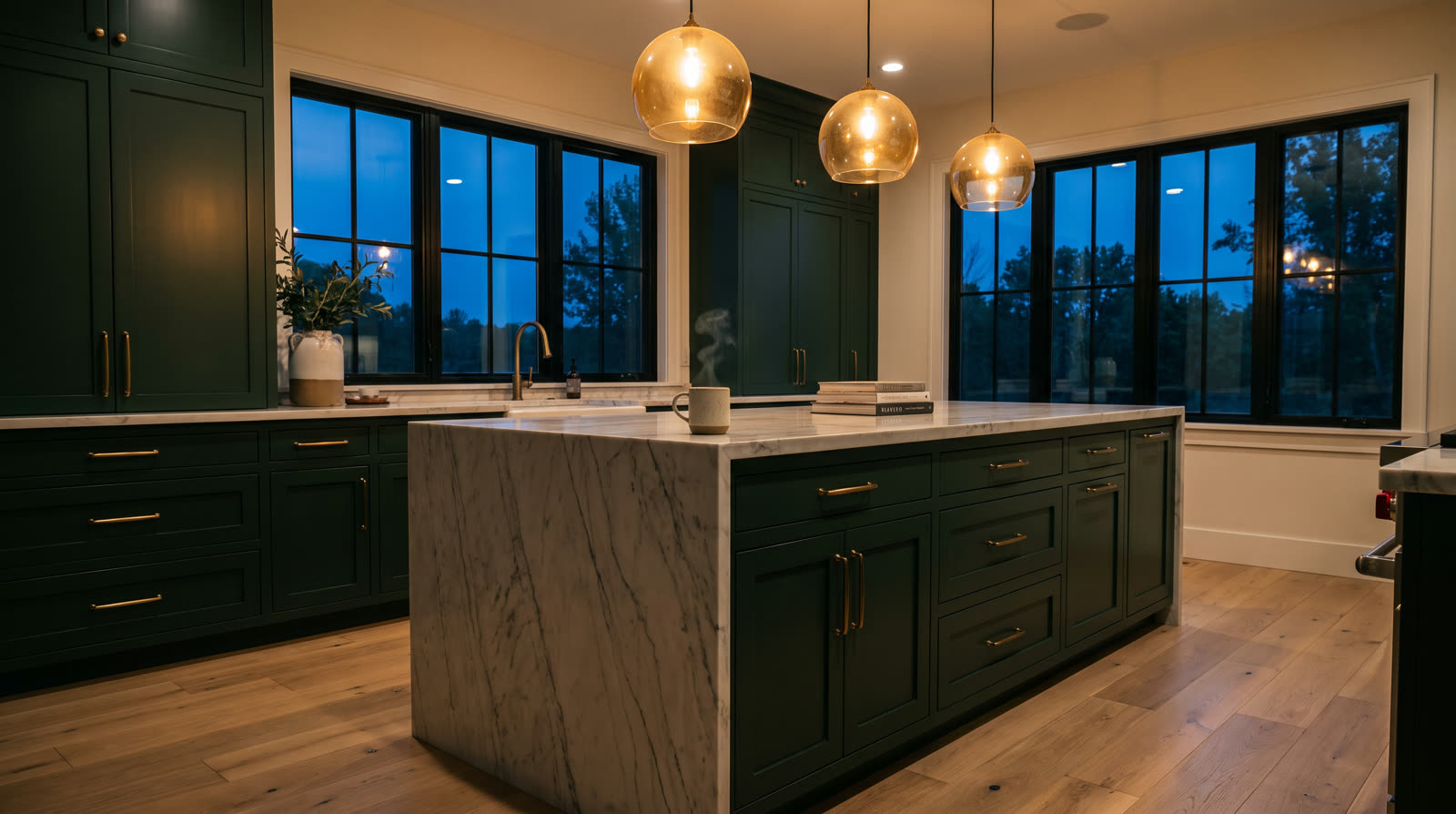 Remodeled kitchen with deep green cabinetry, brass hardware, and a marble waterfall island, lit warmly at dusk